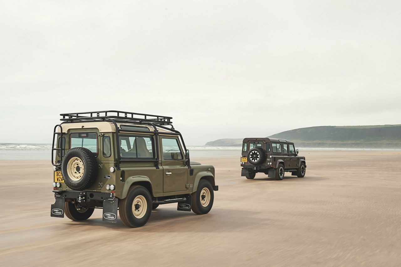Two classic Land Rover Defenders travelling along a wide sandy beach with the sea and distant cliffs behind them.