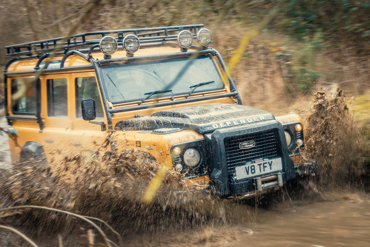 Yellow Land Rover Defender driving off‑road through mud with water splashing across the bonnet.