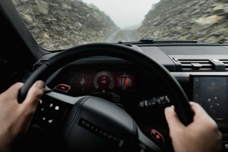 Driver’s-eye view from inside a Defender SUV, showing hands on the steering wheel, digital displays, and a rocky off-road trail ahead.