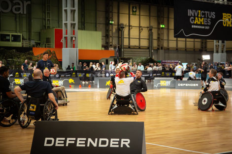 Wheelchair rugby match in an indoor arena, with athletes competing on a wooden court beneath Invictus Games branding and Defender sponsorship signage.