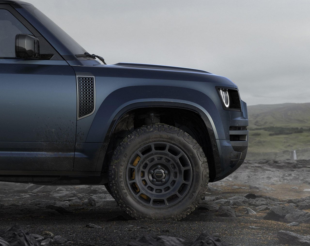 Close-up of a blue off-road SUV’s front wheel and fender, highlighting rugged tires, durable bodywork, and a rocky outdoor landscape.