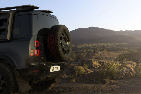 Rear view of a rugged off-road SUV parked on a rocky overlook, with a rear-mounted spare tire and mountains in the background.
