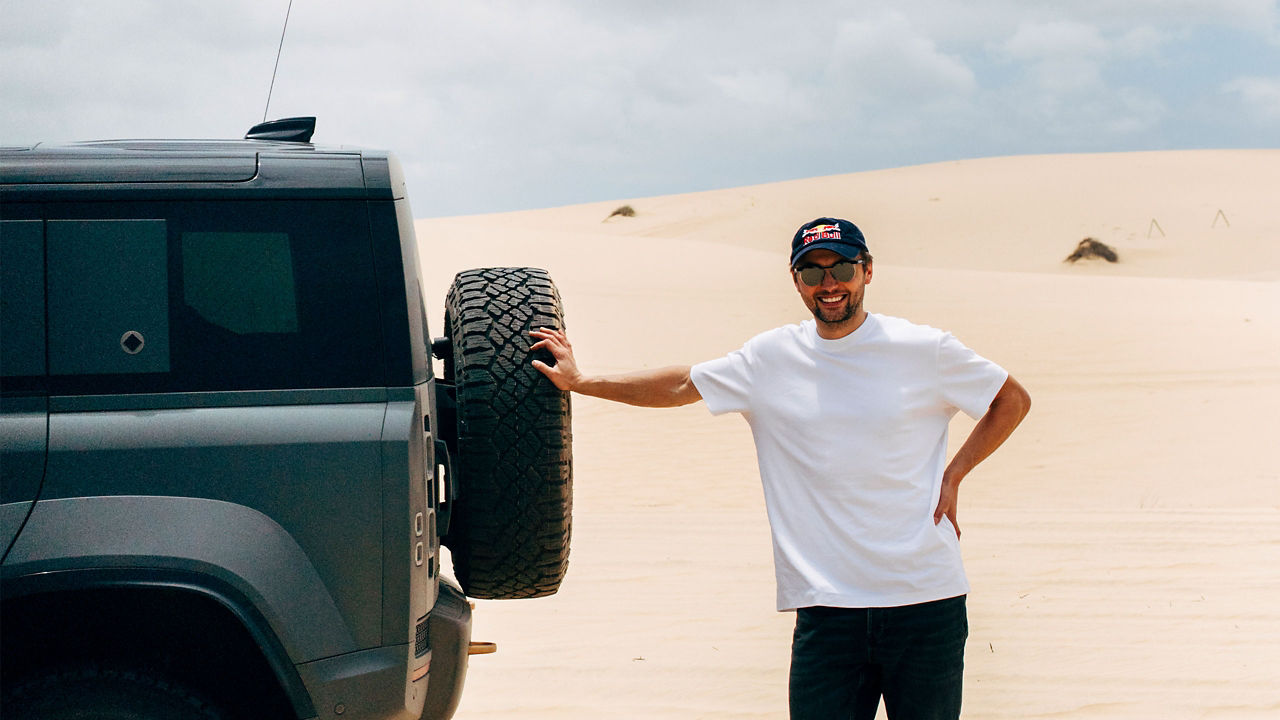 Pilot Andrzej Bargiel leaning on the rear spare wheel of a Defender 110 with the desert dunes in the background.