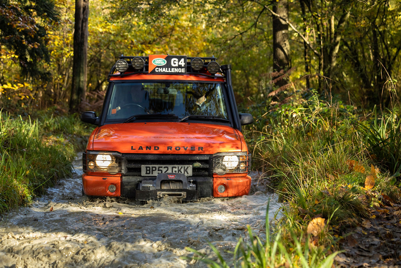 Orange Land Rover Discovery G4 Challenge vehicle with roof‑mounted lights driving through water in a woodland area in autumn.