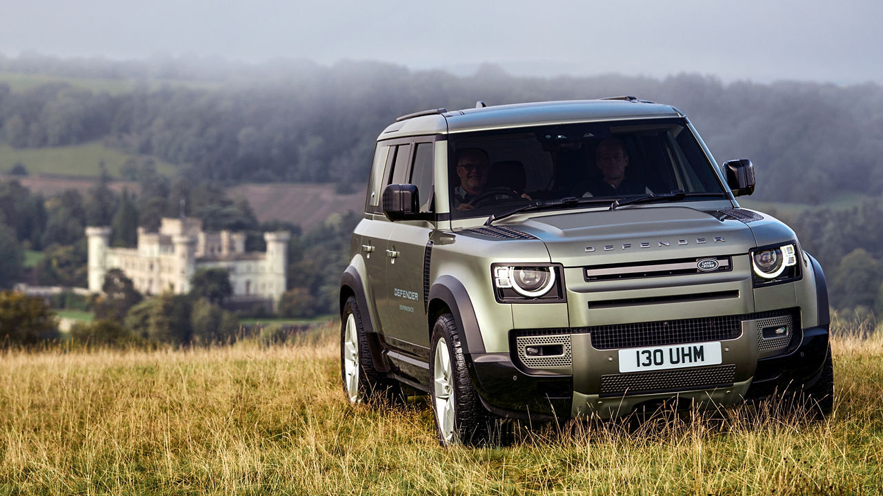 Silver Defender parked on grassy field with hills and buildings in background.