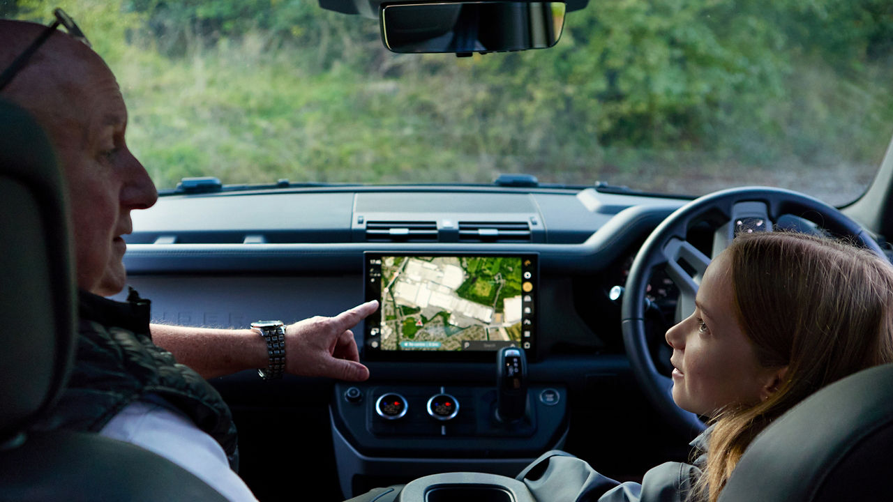 A person in the driver’s seat points at a navigation screen while another person in the passenger seat looks on, inside a Defender.