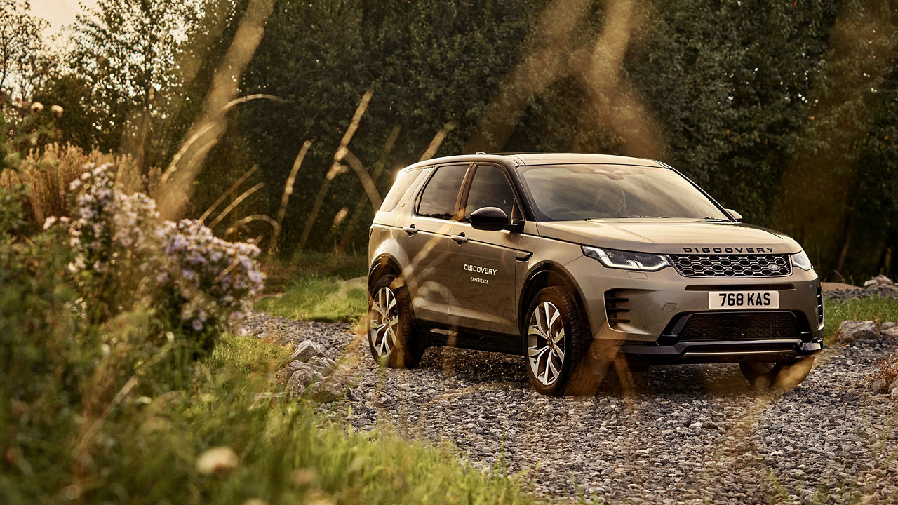 Beige Discovery parked on a gravel path surrounded by grass and trees. 