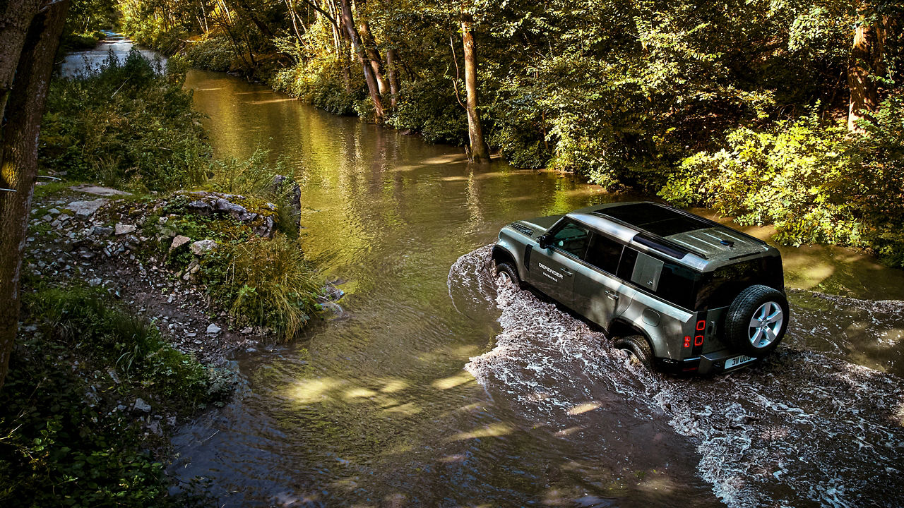 Black Defender crossing a shallow river in a forest.