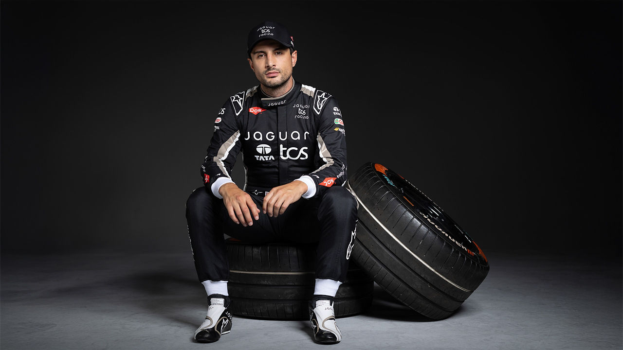 A racing driver in a Jaguar TCS Suit sits on a tire with another tire beside him.