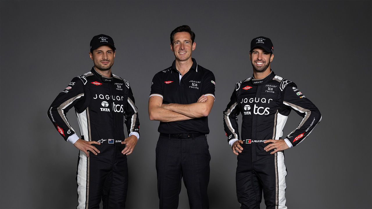 Three men pose in a studio, two in a Jaguar TCS racing suit stand on either side of one in a black team shirt.