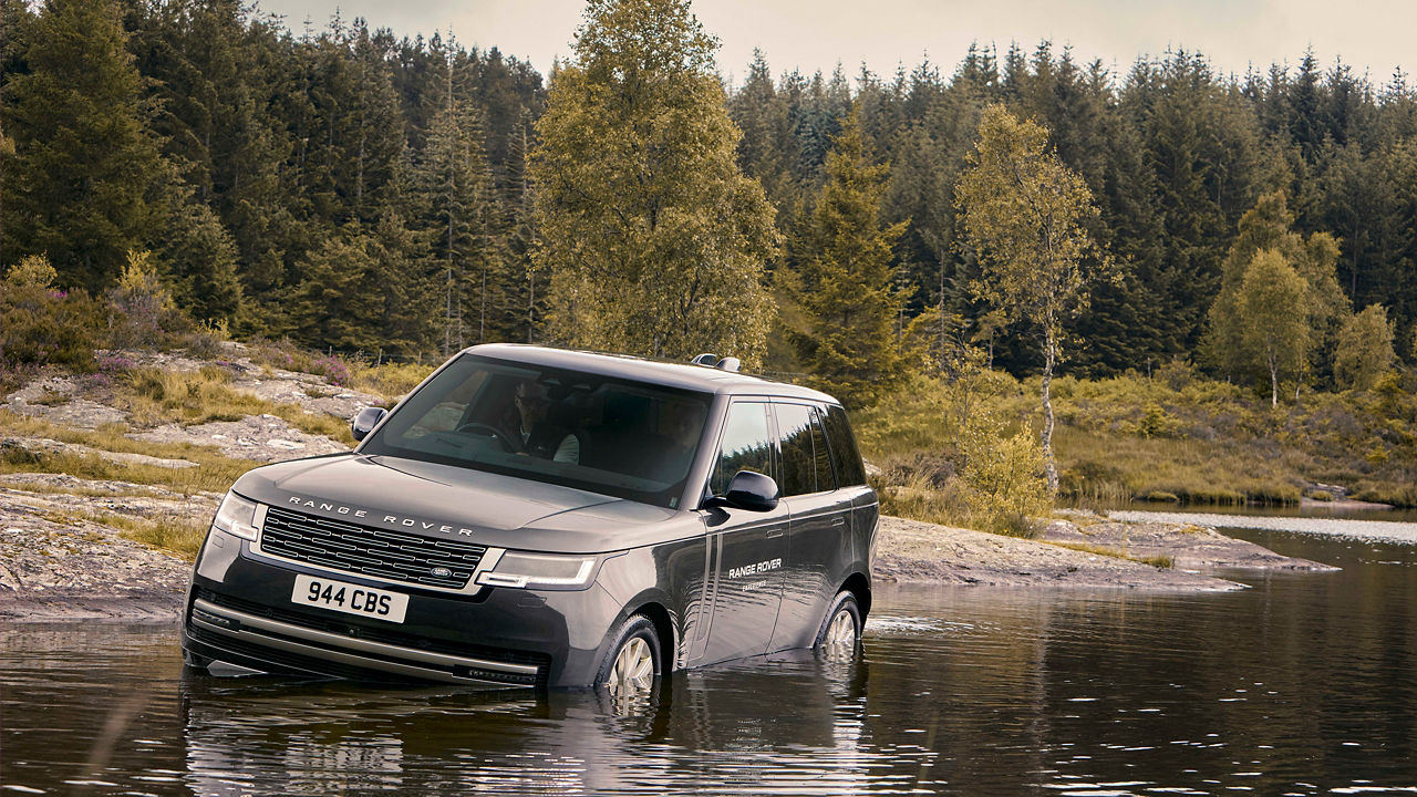 A silver Range Rover driving through shallow water with a forested background.