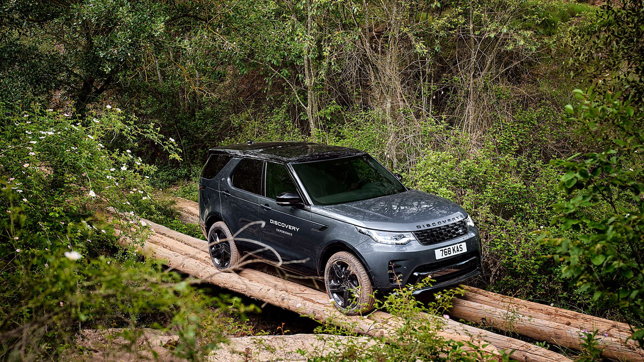 A dark Discovery driving over logs through a dense, green forest.