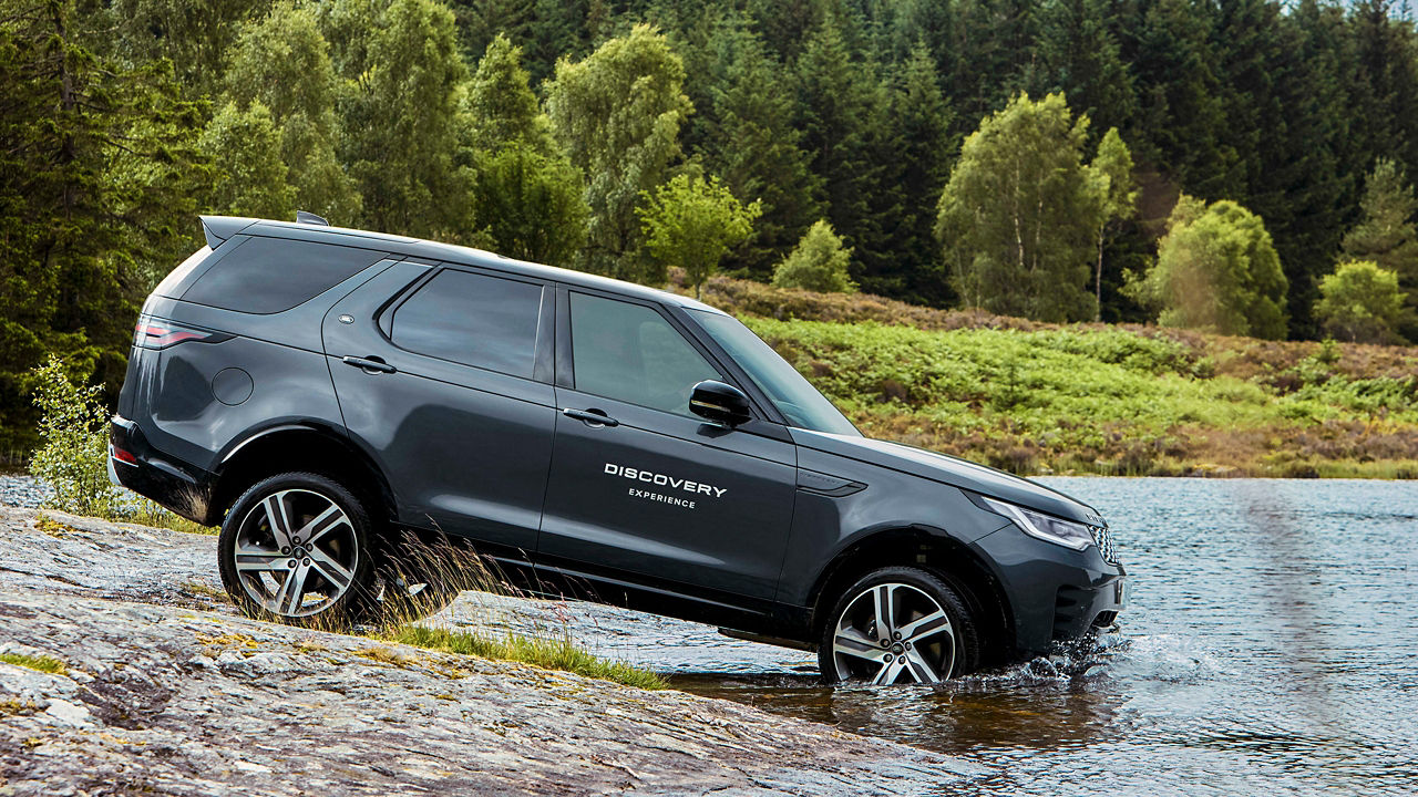 Black Discovery driving through a shallow river with a forest in the background.