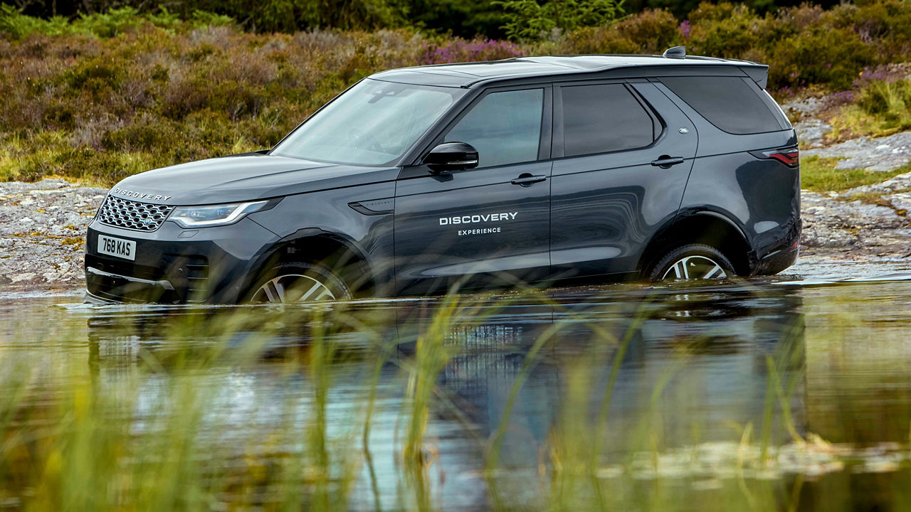 Grey Discovery driving through shallow water in a grassy landscape.