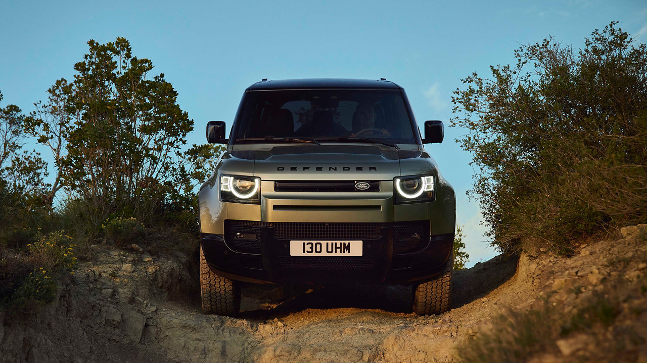 Front view of Defender driving on a dirt trail between trees under a clear sky.
