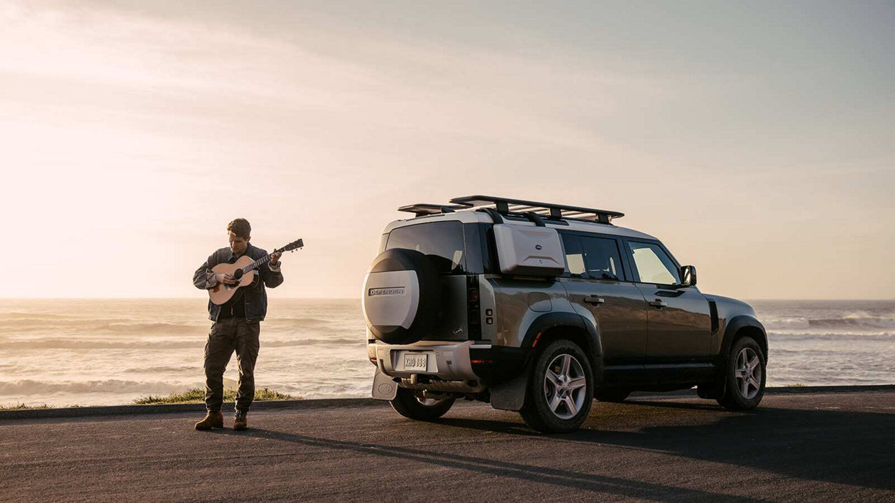 John Mayer playing guitar next to a Land Rover Defender