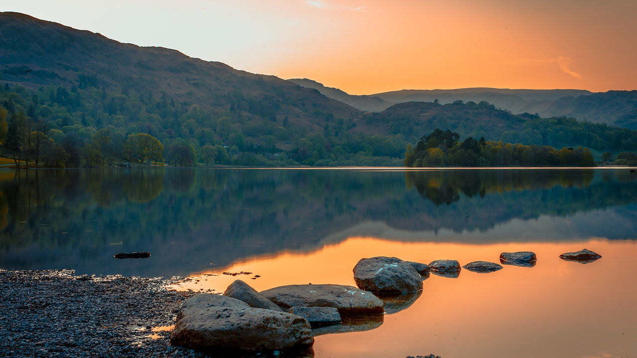 A sunset, grassy mountainside and treeline are reflected in a lake