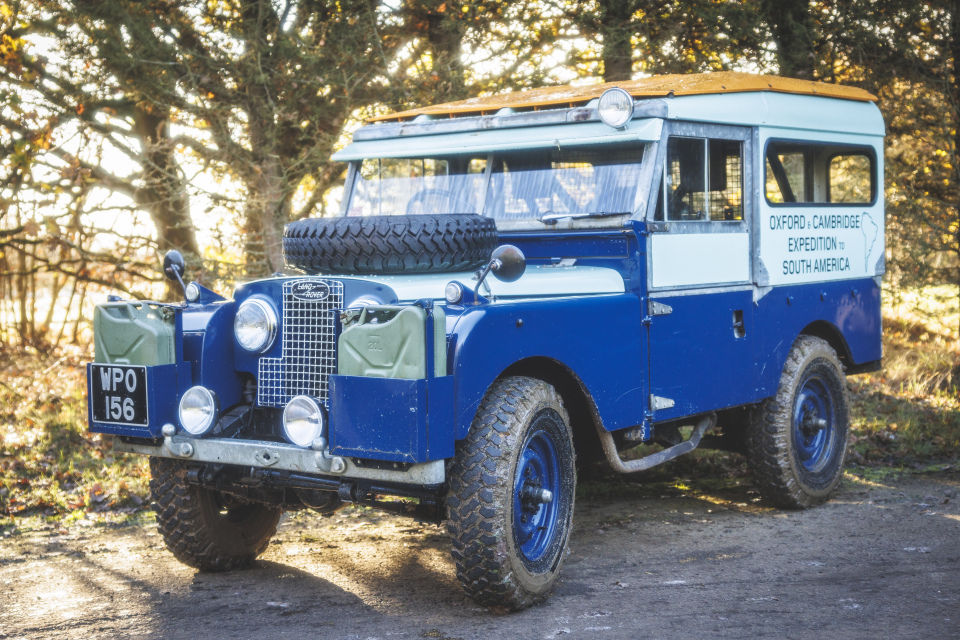 Classic blue and white Land Rover Series 1 parked on a woodland path with sunlight filtering through the trees.