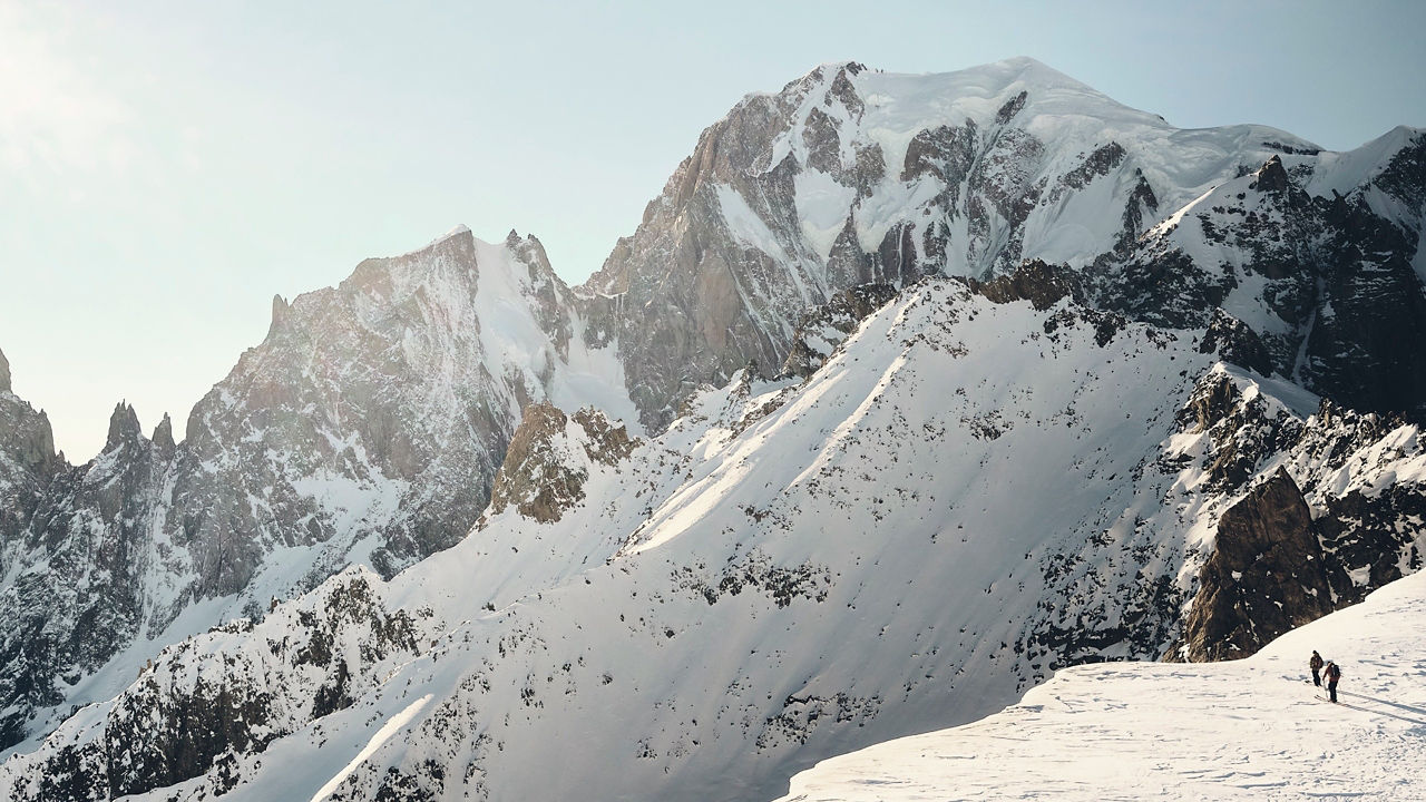 Two individuals hiking a steep mountains edge, surrounded by rocky and snowy mountains and blue sky, with views of the landscape in the background.