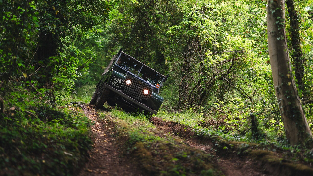 A classic Land Rover with headlights on tackles a rugged, muddy forest trail, tilted on uneven terrain and surrounded by dense green foliage.